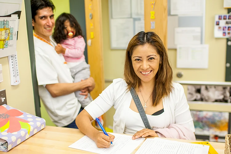 Smiling woman signing her child in at front desk while Dad holds preschool girl behind her.