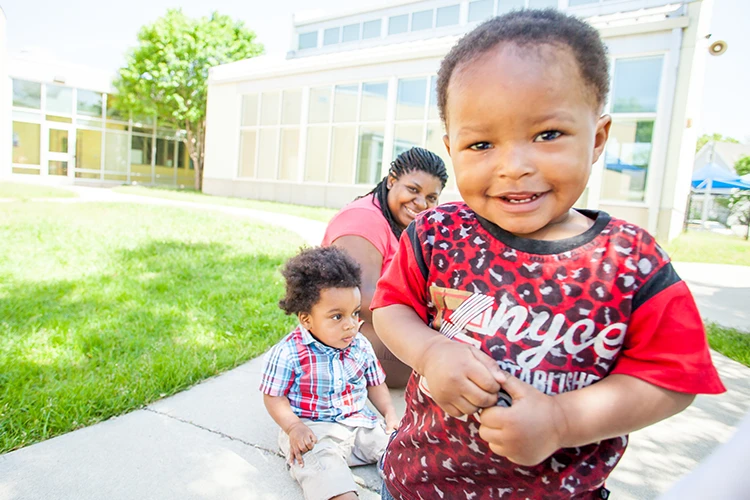 Close up of toddler boy with woman and another toddler in background outside of an Educare school.